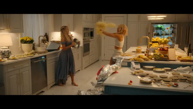 Emily Alyn Lind standing barefoot in a kitchen in a blue dress in a scene.