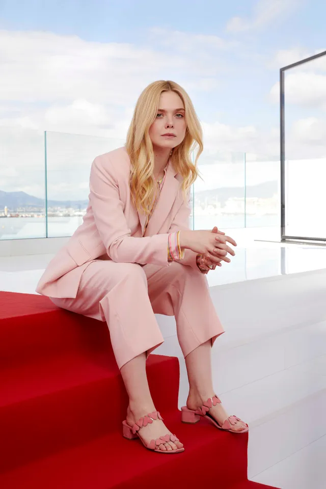 Elle Fanning sitting in a pink suit and pink heeled sandals.