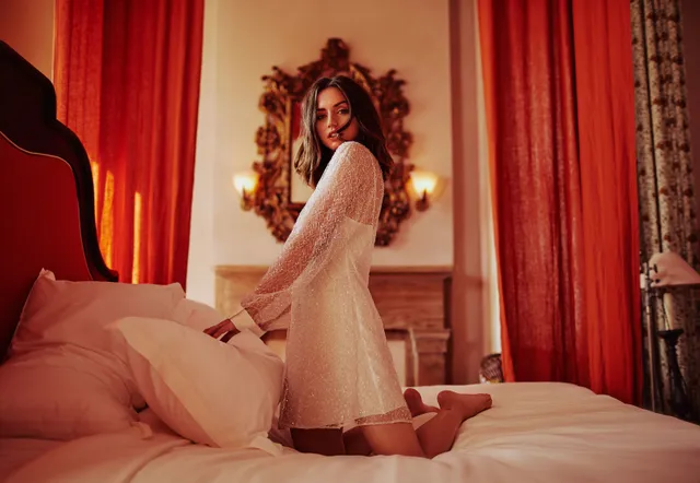 Ana de Armas kneeling on a bed with her bare feet lifted behind her.