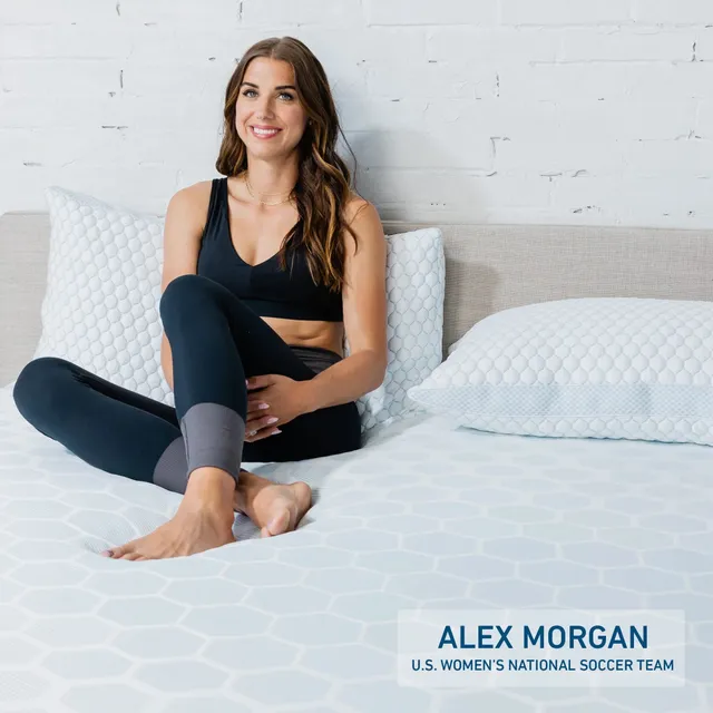 Alex Morgan sitting cross-legged and barefoot on a mattress in a dark exercise outfit.
