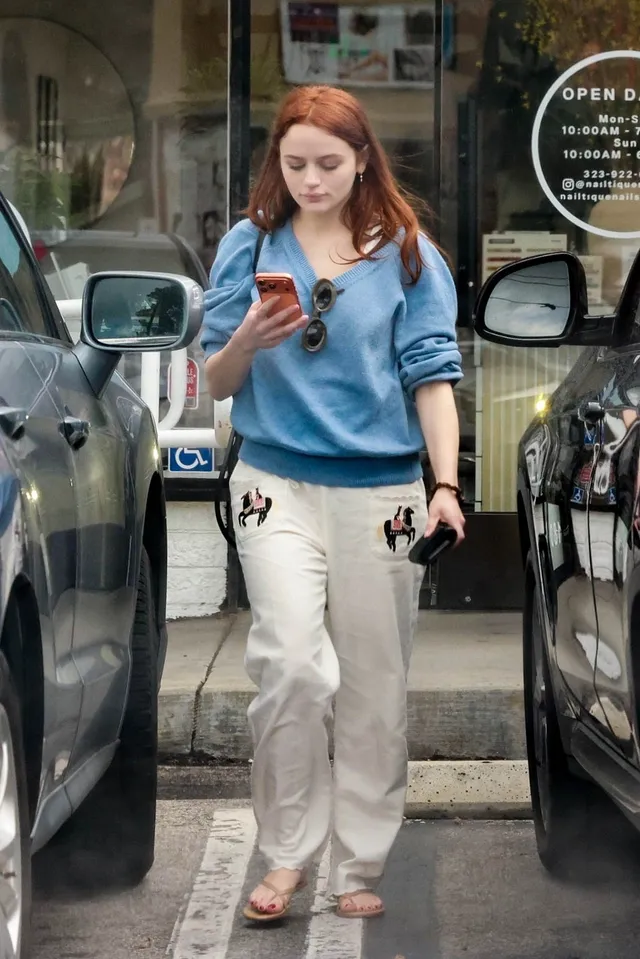 Joey King looking at her phone outdoors in pale trousers and sandals.