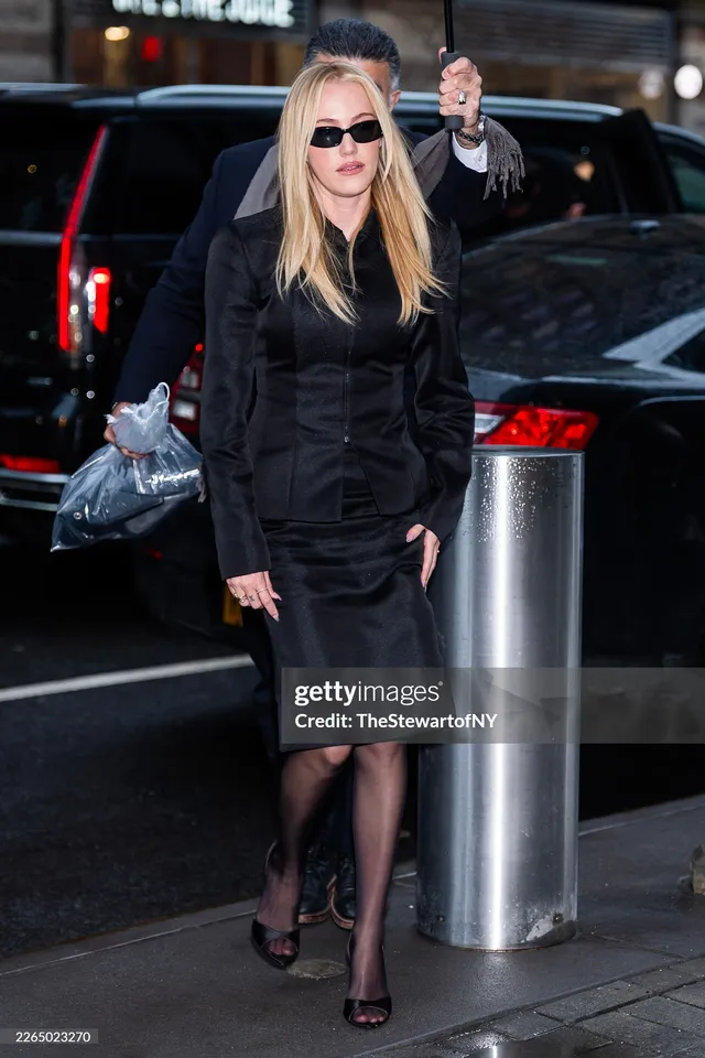 Maika Monroe walking in a black outfit and heels outdoors.