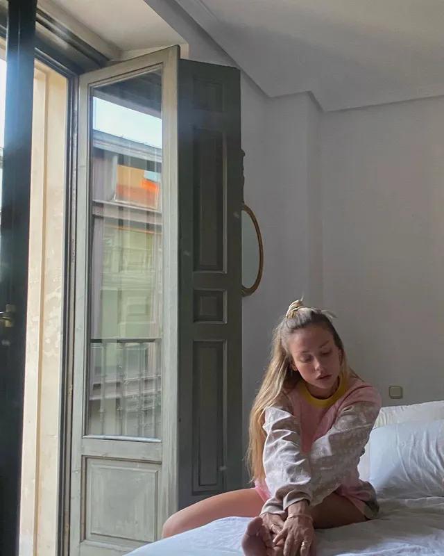 Ester Expósito sitting barefoot on a bed in a pale embroidered shirt.