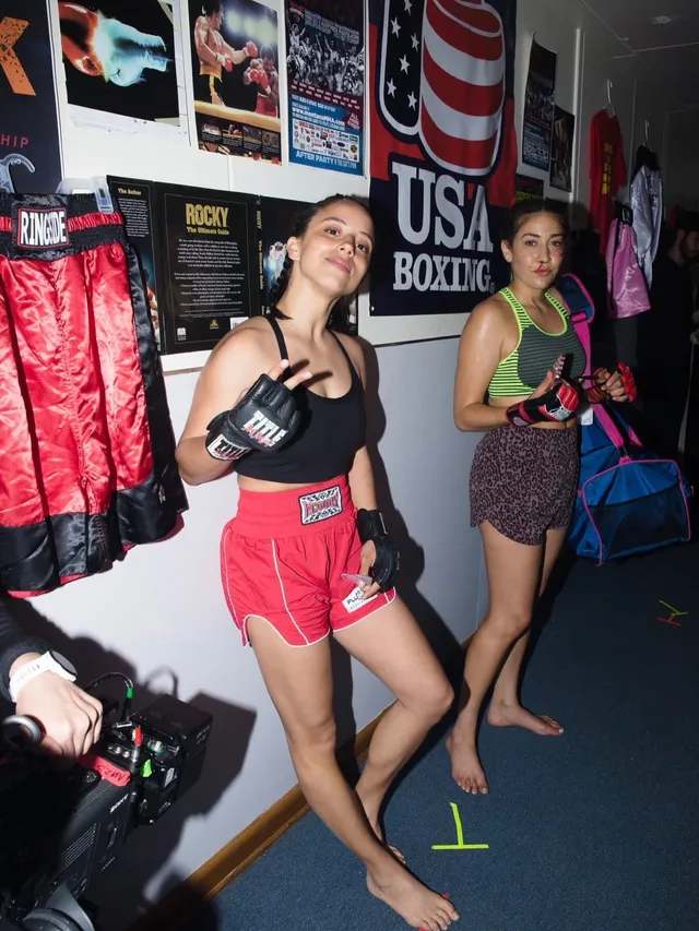 Kiana Madeira standing barefoot in a boxing gym in red shorts and gloves.