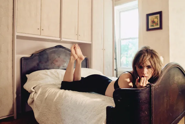 Suki Waterhouse lying on a bed in a black outfit.