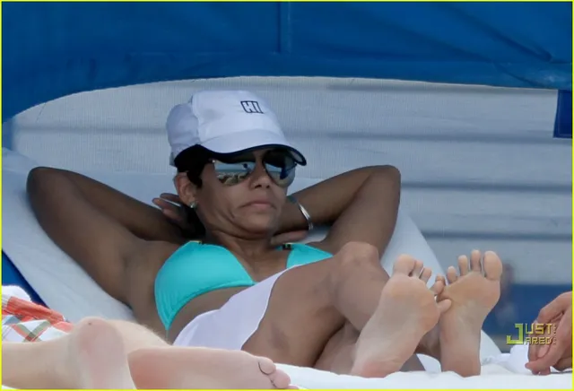 Halle Berry reclining barefoot beside a pool in sunglasses.