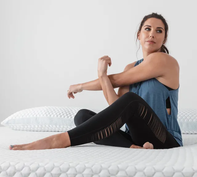 Alex Morgan stretching barefoot on a mattress in a dark exercise outfit.
