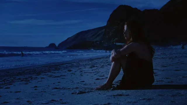Mackenzie Ziegler sitting barefoot on beach sand in a dark scene.