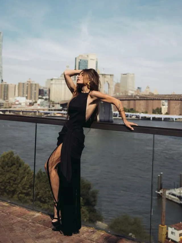 Sarah Shahi posing on a rooftop terrace in a long black dress.