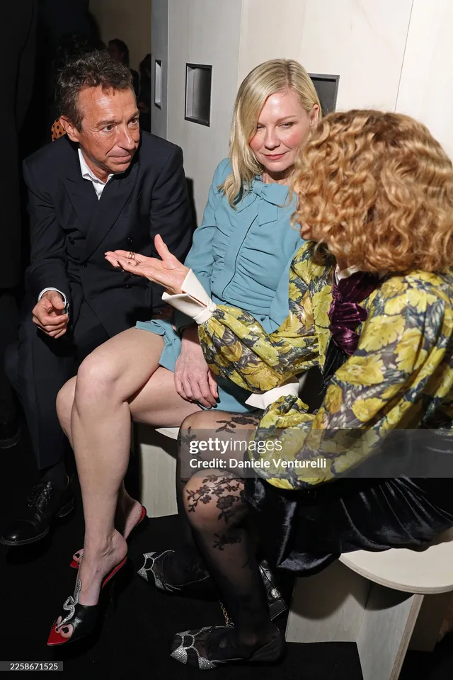 Kirsten Dunst sitting in a blue dress and patterned heels while talking with guests.