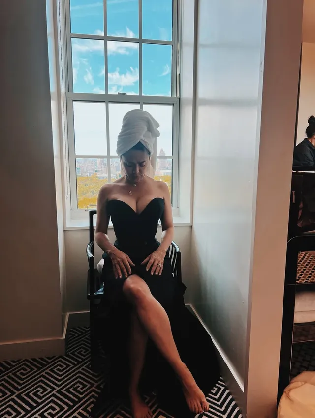 Salma Hayek seated by a window in a black evening gown.
