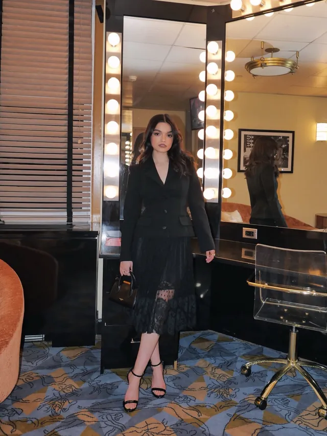 Rachel Zegler standing by a dressing-room mirror in black heels.