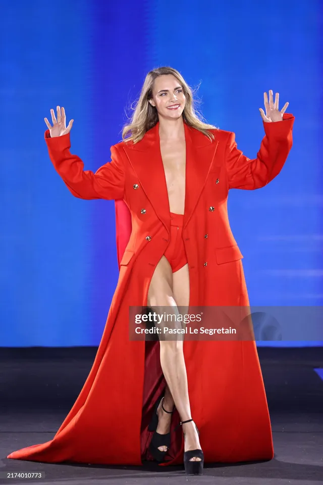 Cara Delevingne waving on a runway in a long red coat.