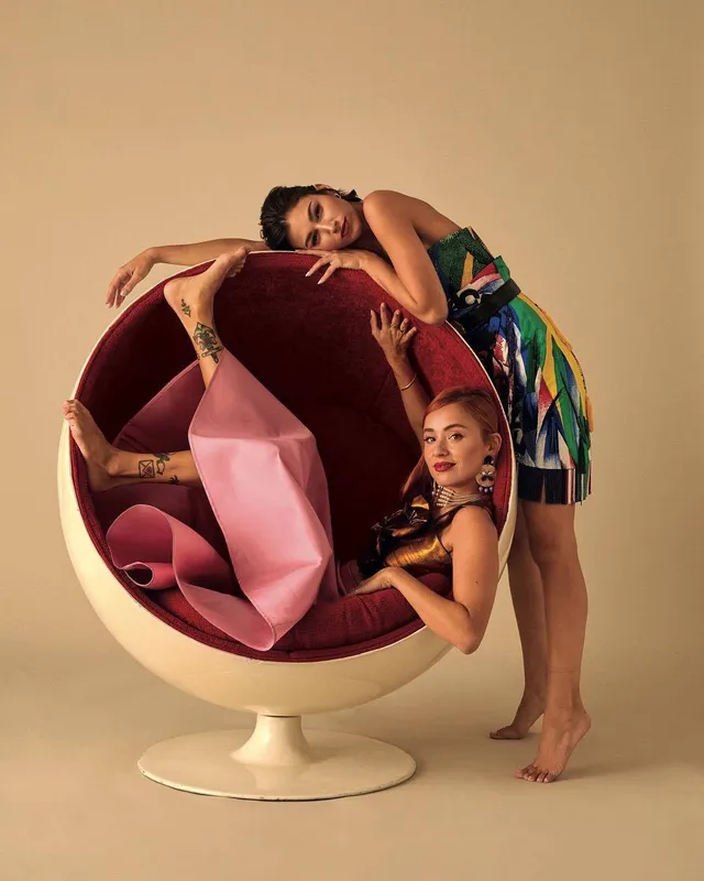 Úrsula Corberó posing barefoot beside a guest and a round chair for a studio portrait.