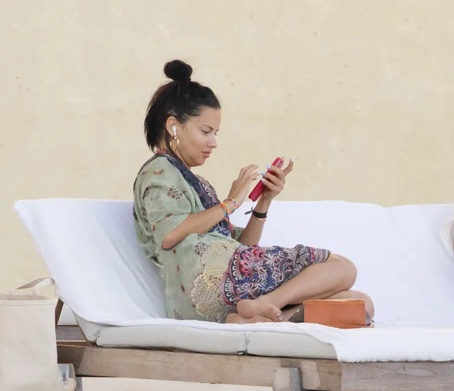 Adriana Lima sitting barefoot on a sun lounger in a patterned dress.