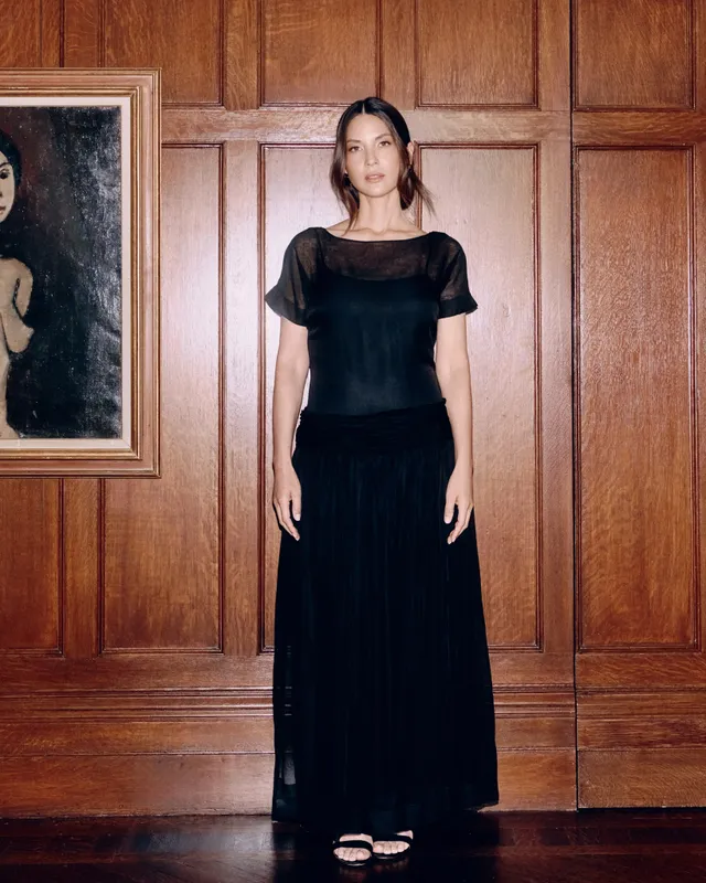 Olivia Munn standing by a wooden wall in a long black dress and heels.