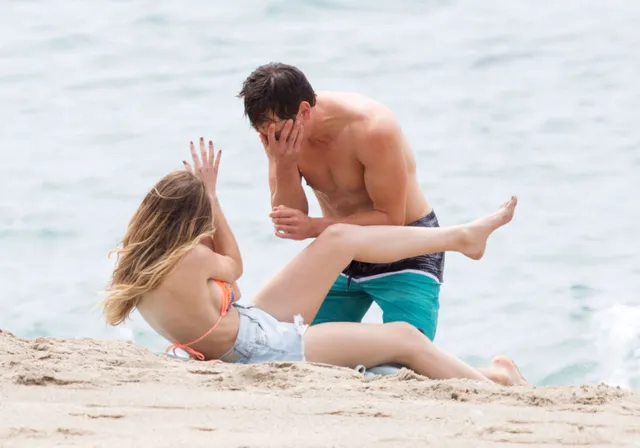 Halston Sage sitting barefoot on a beach beside a man.