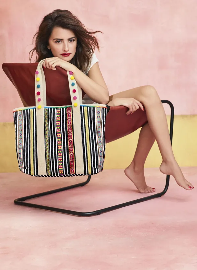 Penélope Cruz sitting barefoot in a red chair beside a colorful bag.