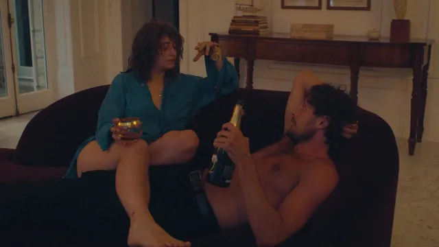 Odessa A'zion sitting barefoot on a sofa with a companion in a scene.