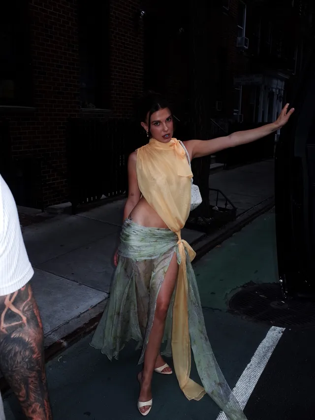 Millie Bobby Brown posing outside in a pale top and patterned skirt with heels.