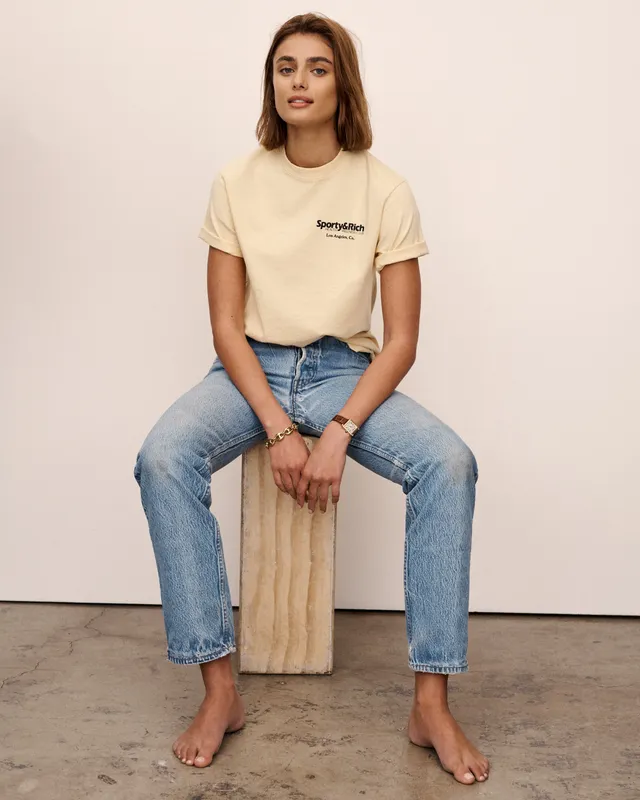 Taylor Hill sitting barefoot on a stool wearing jeans and a white T-shirt.