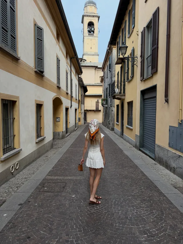 Eugenie Bouchard standing in heeled sandals and a short white dress on a narrow street.