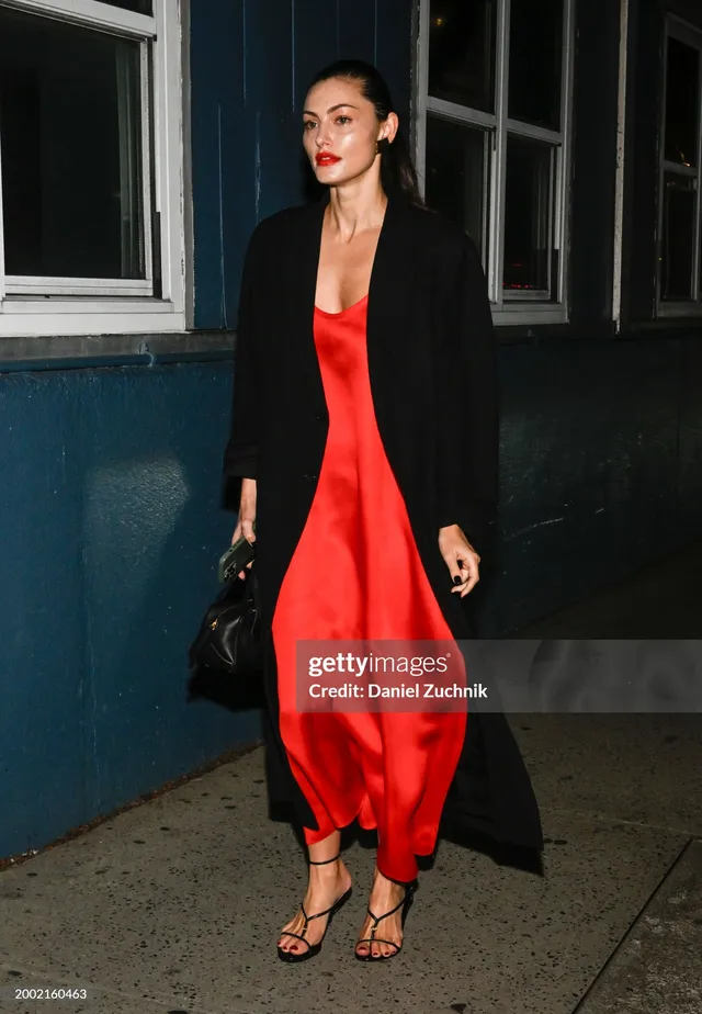 Phoebe Tonkin walking outdoors in a red dress and dark coat.