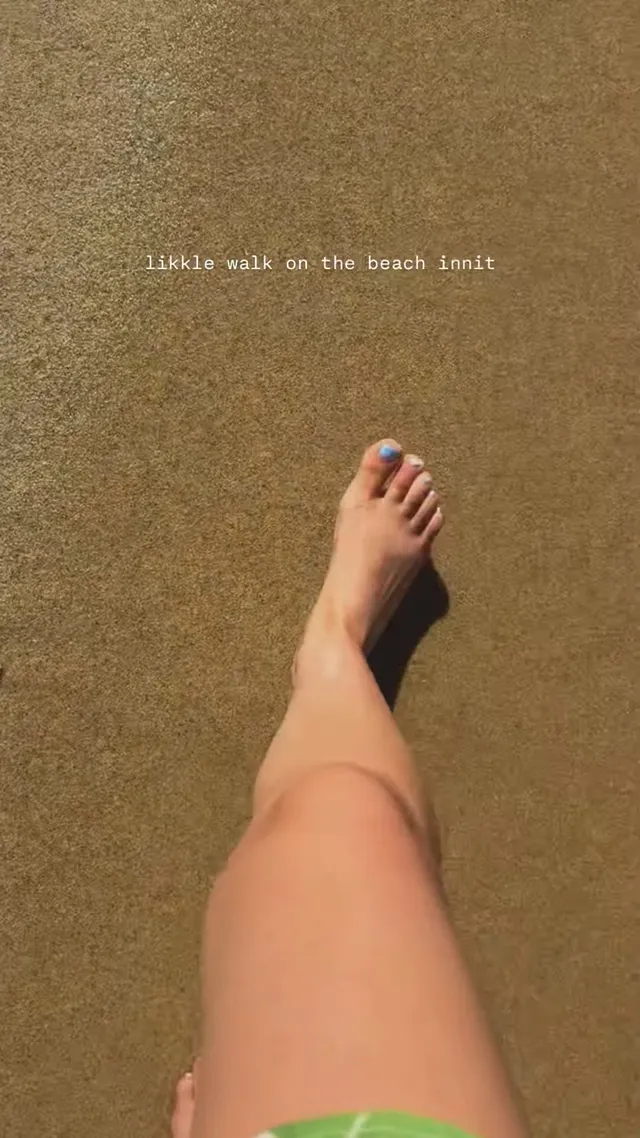 Rachel Zegler stepping barefoot across wet beach sand.