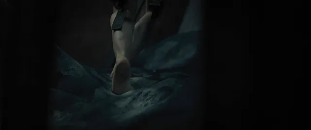 Cailee Spaeny standing barefoot on a metal floor in a dimly lit film scene.