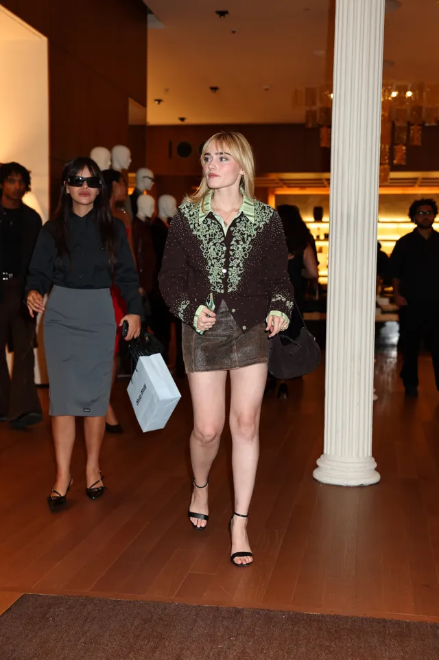 Meg Donnelly walking indoors in a patterned blouse, brown skirt and black heels.