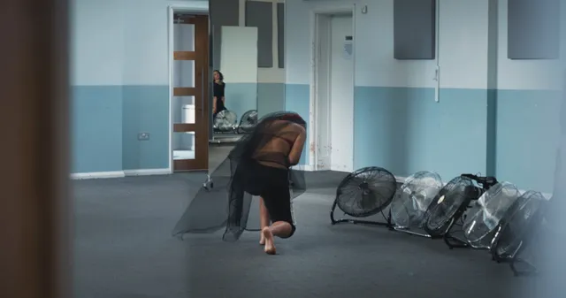 Naomi Scott kneeling barefoot beside floor fans in a rehearsal room.