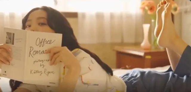 Jisoo lying on a bed in jeans and a pale top with a bare foot raised.