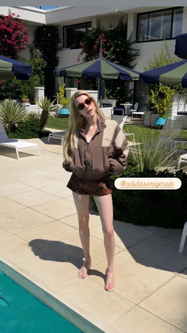 Emma Roberts standing barefoot beside a pool in a gray jacket and black shorts.