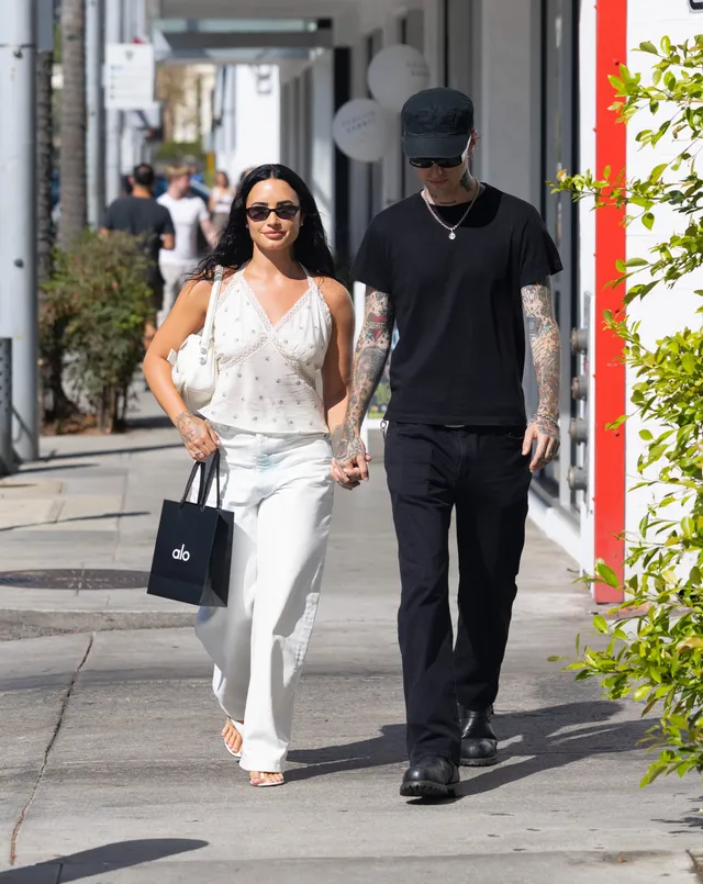 Demi Lovato walking beside another person in white trousers and flat sandals.