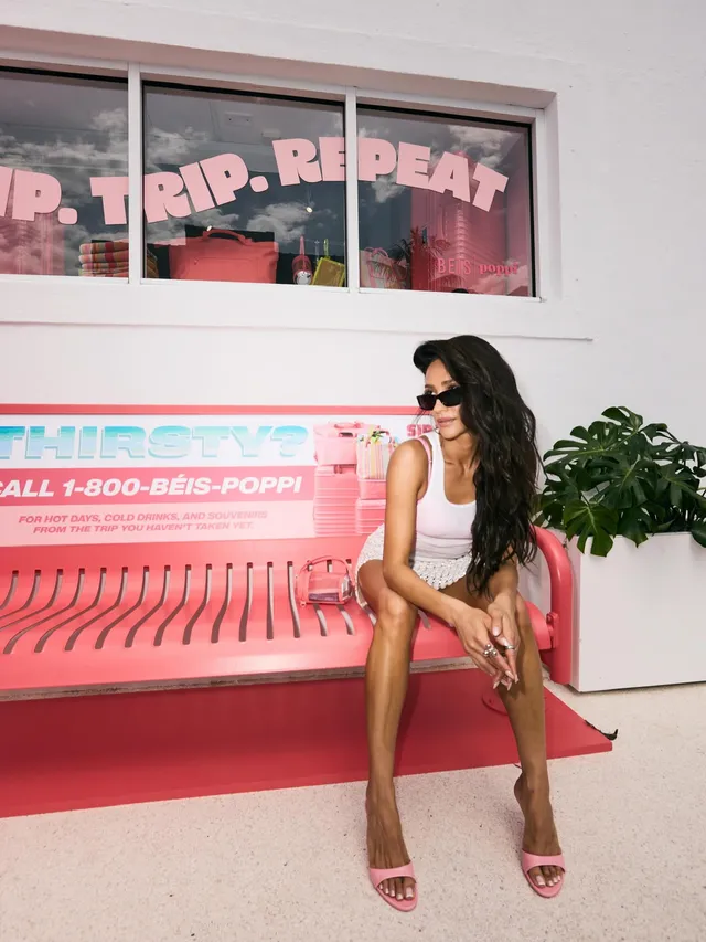 Shay Mitchell sitting beside a pink display in pink heeled sandals.