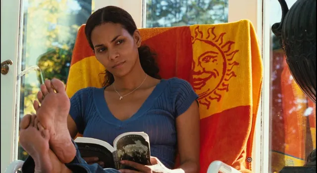 Halle Berry sitting barefoot in a blue top with a book.