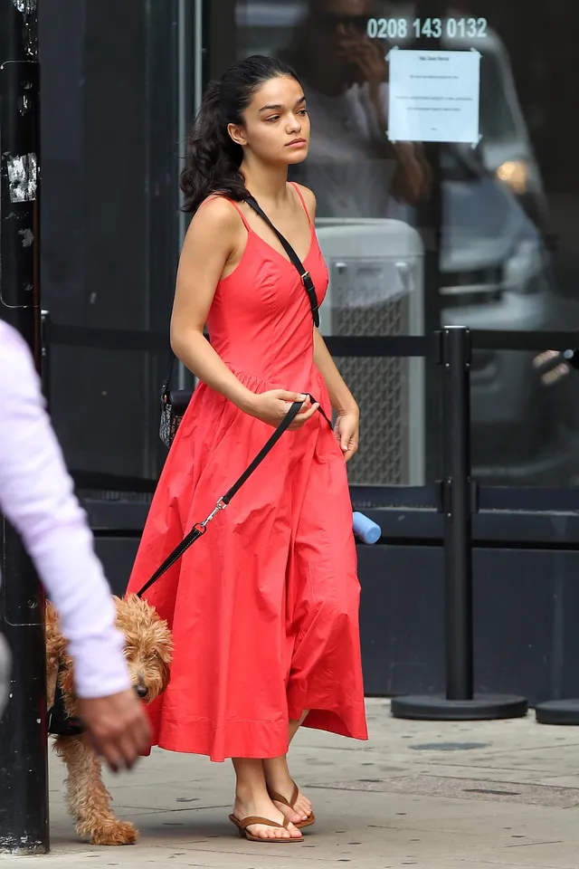 Rachel Zegler standing outdoors with a dog in a red dress and flat sandals.