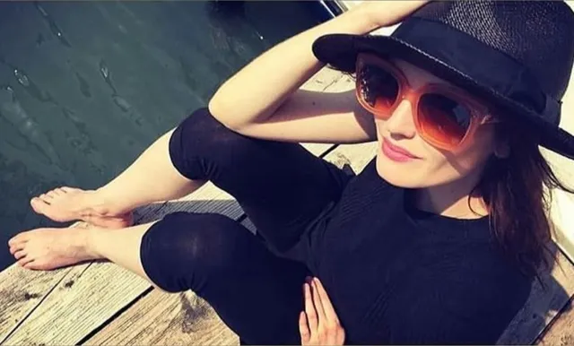 Eva Green sitting barefoot on a wooden surface wearing a hat.