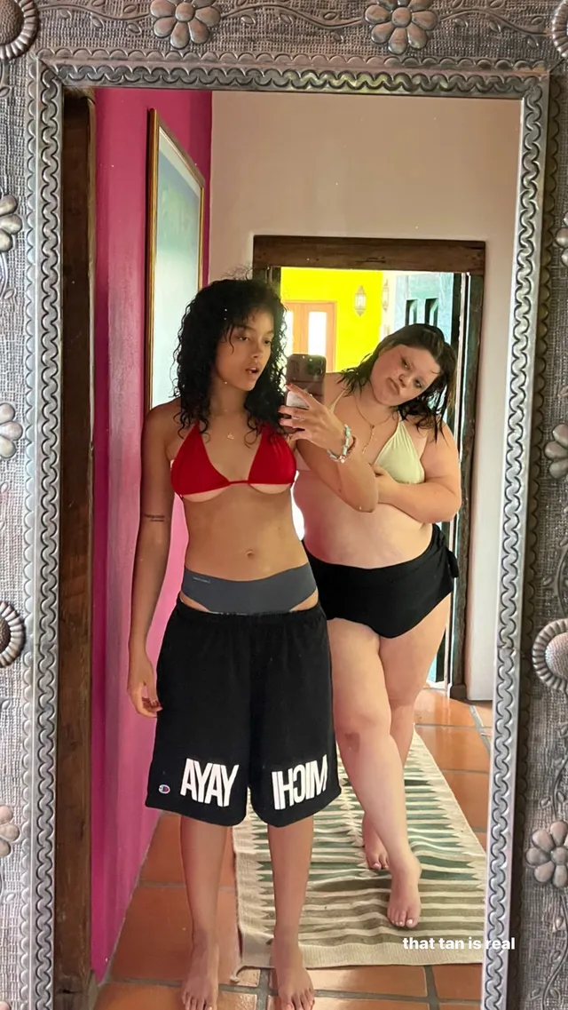 Whitney Peak taking a mirror selfie with a friend in a red bikini top, shorts and flat sandals.