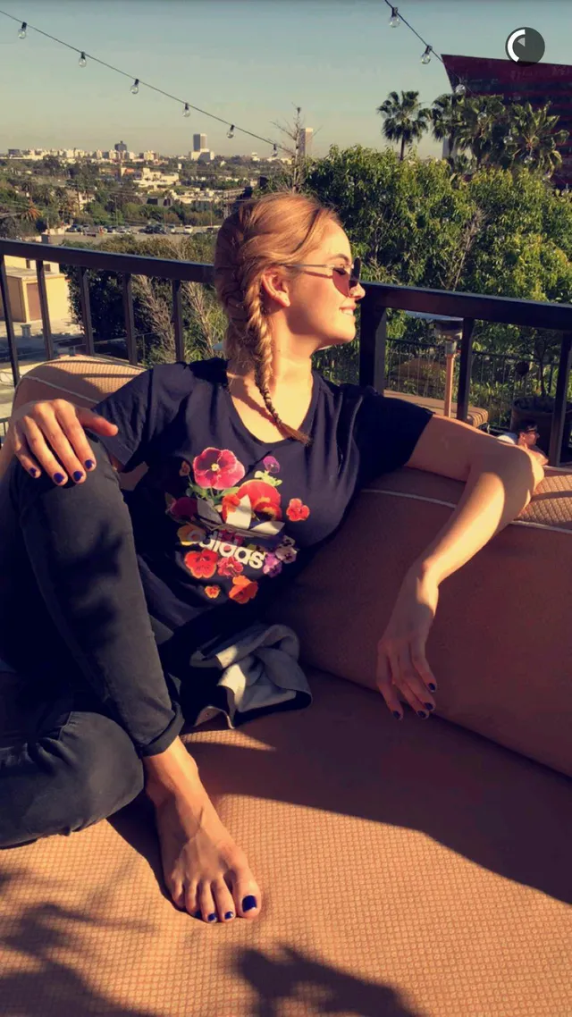 Debby Ryan sitting barefoot on an outdoor seat in a dark T-shirt.