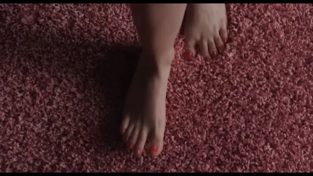 Cailee Spaeny standing barefoot on a patterned bedroom carpet.