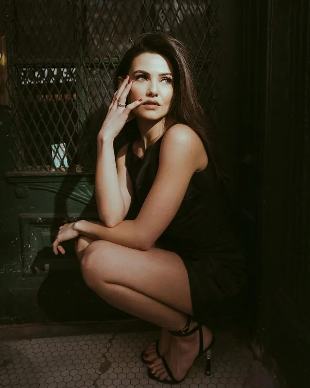 Danielle Campbell crouching beside a gate in a black dress.