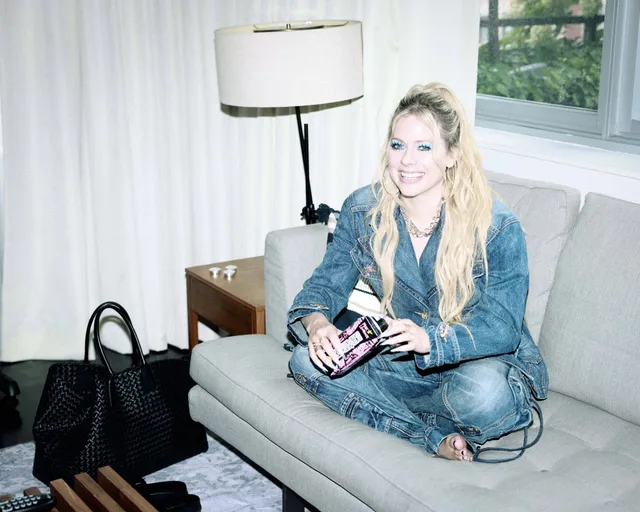 Avril Lavigne sitting barefoot on a pale sofa in a denim outfit.