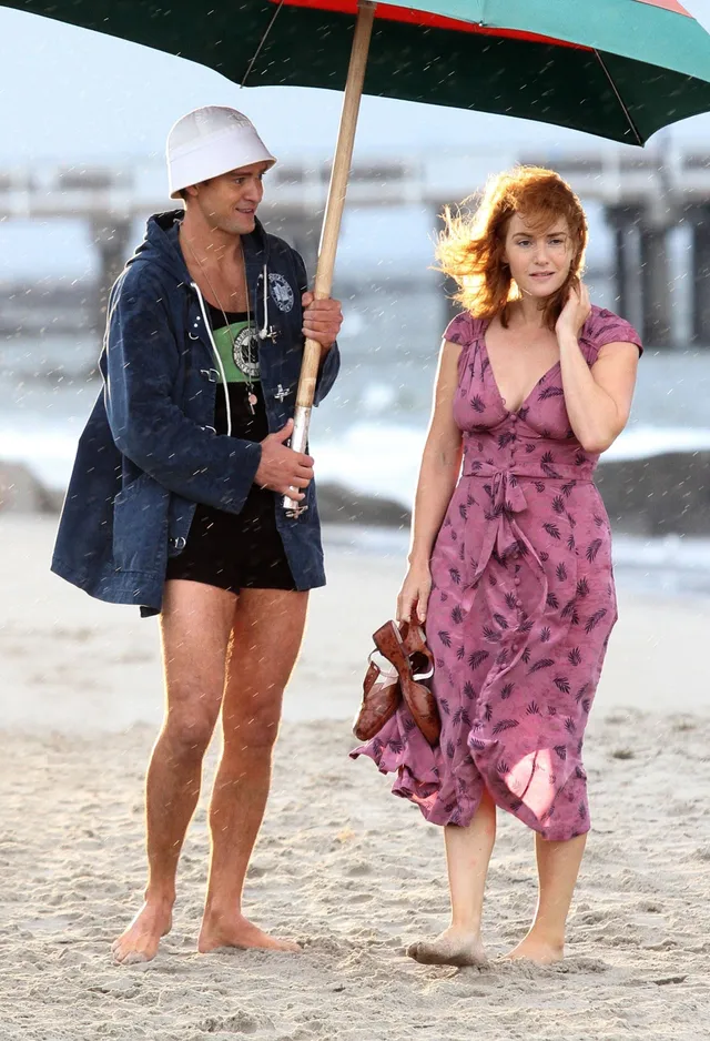 Kate Winslet walking barefoot on a beach in a pink dress beside a companion.