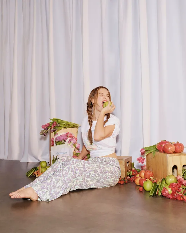 Millie Bobby Brown: seated barefoot in a floral dress beside flowers and fruit.
