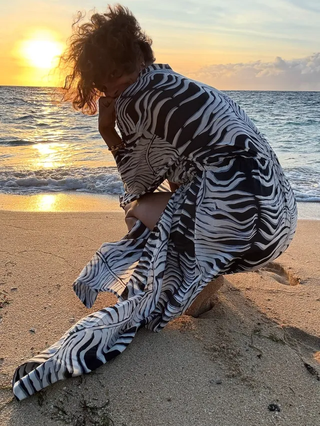 Halle Berry kneeling barefoot on a beach in a striped cover-up.