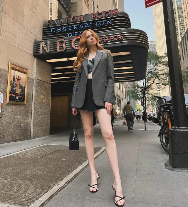 Karen Gillan posing outside a cinema in a gray jacket and heels.