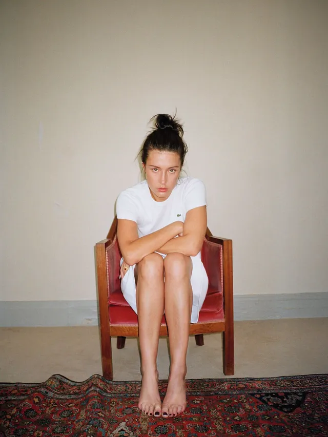 Adèle Exarchopoulos sitting in a wooden chair in a white shirt.