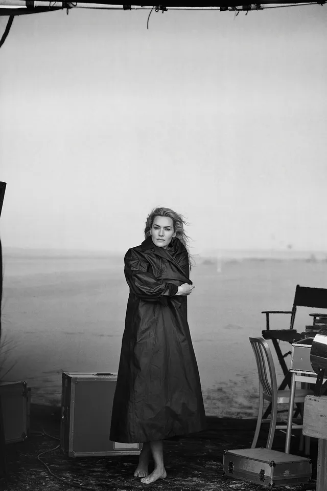 Kate Winslet standing barefoot in a black dress indoors.