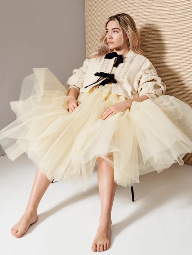 Florence Pugh sitting barefoot in a pale full skirt for a studio portrait.